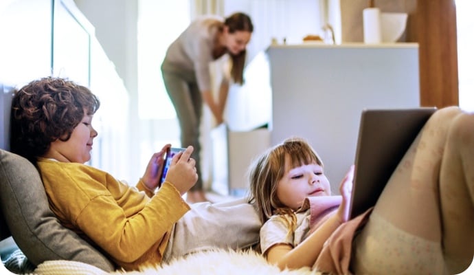 Niños recostados usan tablets mientras la madre en el fondo organiza la cocina.